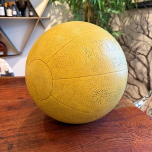 May include: A yellow medicine ball with a textured surface and segmented design. The ball is round and appears to be made of rubber or a similar material. The ball is on a wooden surface.