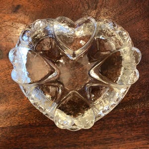 Puede incluir: Plato de servir transparente en forma de corazón. El plato presenta seis compartimentos en forma de corazón alrededor de un área central en forma de corazón. El vidrio tiene una superficie texturizada y el plato está sobre una superficie de madera oscura.