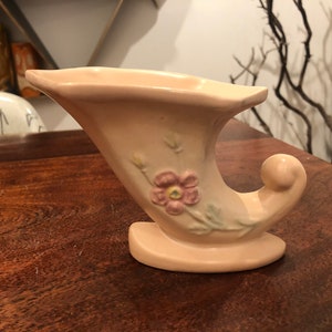 Może przedstawiać: Różowy ceramiczny wazon w kształcie rogu z kwiatowym wzorem. Wazon ma różowy kwiat z żółtymi akcentami i zielonymi liśćmi.