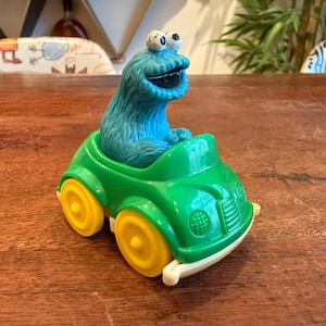 May include: A Cookie Monster toy figure in a green car with yellow wheels. The Cookie Monster figure is blue with white eyes and a large open mouth. The car has a white bumper and is sitting on a wooden surface.