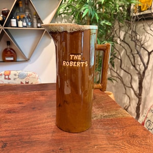 May include: A brown ceramic pitcher with a handle and a spout. The pitcher has the words "THE ROBERTS" embossed on the front. The top rim has a light brown and beige glaze. The pitcher is sitting on a wooden table.