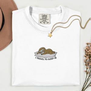 Könnte beinhalten: Weißes Comfort Colors T-Shirt mit einem aufgestickten Faultier-Motiv auf der Vorderseite. Das Faultier liegt auf einem Kissen, darunter der Text "I TRAVEL TO SLEEP IN". Eine goldene Stern-Anhänger-Halskette liegt auf dem Shirt.