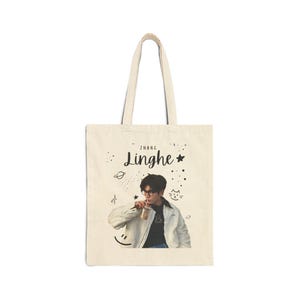 May include: A beige canvas tote bag featuring a printed photograph of a person wearing glasses and a white jacket while holding a drink. The text Zhang Linghe is printed above the image, surrounded by small black hand-drawn doodles of stars.