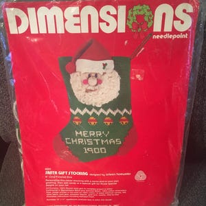 May include: A Christmas needlepoint kit featuring a Santa Claus stocking design. The kit includes a green stocking with a Santa face, the words "MERRY CHRISTMAS 1900", and the Dimensions needlepoint logo on a red background.