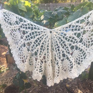 May include: A white crocheted shawl with a delicate lace pattern. The shawl is hanging on a clothesline outdoors.