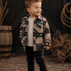 May include: A young child wearing a patterned jacket with brown, teal, and white geometric designs. The child is also wearing a white thermal shirt, black pants, and brown boots. The background includes a wooden door and a barrel.