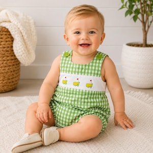 May include: A baby wearing a green and white gingham romper with embroidered golf course flags. The romper has green buttons on the shoulders and is paired with white shoes. The baby is sitting on a white rug.