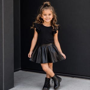 May include: A black ribbed short-sleeve top with ruffled shoulders paired with a black faux leather pleated skirt. The outfit is completed with black lace-up ankle boots. The ensemble features a consistent monochromatic black color palette.
