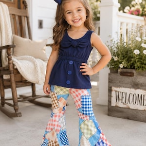 May include: A young girl wearing a navy blue top with a bow and three buttons, paired with patchwork bell-bottom pants. She also wears a matching bow in her hair and white sandals. The outfit is a mix of patterns and colors.