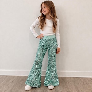 May include: A young person wearing a white long-sleeved shirt and mint green sequin bell-bottom pants. The pants have a mint green waistband. The person is wearing white sneakers. The background is a plain white wall.