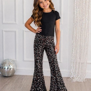 May include: A young person wearing a black short-sleeved top and flared sequin pants. The pants are black with a pattern of silver and copper sequins. The person is smiling and posing.