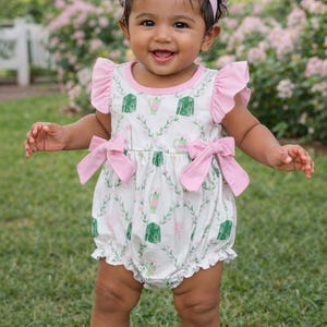 May include: A baby wearing a white romper with a green and pink floral pattern, pink bows, and a matching headband. The romper has ruffled sleeves and leg openings. The baby is smiling and standing on green grass.