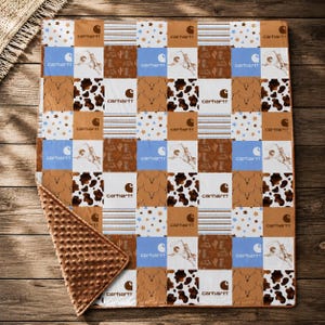 May include: A patchwork blanket featuring a repeating pattern of brown, white, and light blue squares. The design includes the Carhartt logo, illustrations of cowboys on bucking horses, cow print, western boots, cacti, and small star motifs.