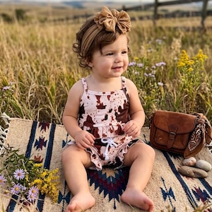 May include: A baby wearing a cow print romper and a matching headband sits on a patterned blanket. A brown leather purse and a bandana are next to the baby. The background is a field with tall grass and a wooden fence.