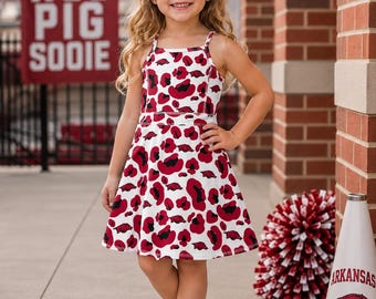Peut inclure: Une robe sans manches avec une base blanche ornée d'un motif répétitif de taches léopard rouges et noires ainsi que de petites silhouettes de cochons rouges. La robe possède de fines bretelles et une jupe évasée. Un grand nœud rouge foncé est inclus.