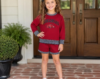 Peut inclure: Un ensemble composé d'un sweat-shirt à manches longues et d'un short bordeaux, agrémenté d'un liseré à carreaux vichy noir et blanc au col, aux poignets et à l'ourlet. Le sweat-shirt porte le mot Arkansas en noir au-dessus d'un motif rouge et blanc.