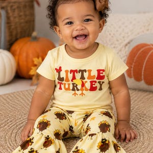 May include: A light yellow short-sleeved t-shirt with the text Little Turkey in brown, orange, and red, paired with matching light yellow bell-bottom pants featuring an all-over print of brown and yellow turkeys and small orange leaves.