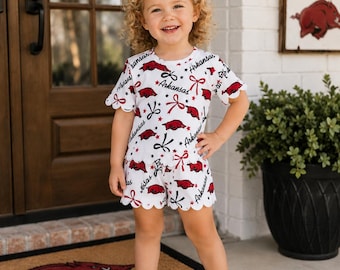 Peut inclure: Un ensemble blanc composé d'un haut à manches courtes et d'un short, orné d'un motif répétitif de logos razorback rouges de l'Arkansas, de texte noir écrit Arkansas et de nœuds rouges. Les manches et l'ourlet du short ont une finition festonnée.