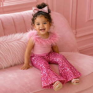 Op de afbeelding: Een kind draagt een roze outfit met een gevederde top, een broek met pailletten en een strik in het haar. Het kind zit op een roze bank en lacht.