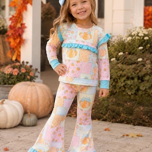May include: A long-sleeve top and bell-bottom pants set featuring a patchwork pattern of pumpkins, orange gingham, and floral prints in pastel pink, blue, and orange. The top has a ruffled chest detail, and both pieces have light blue ruffled hems.