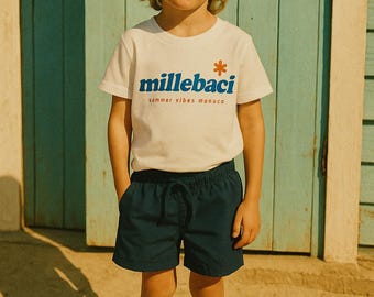 May include: A child wearing a white t-shirt with the text "millebaci sommer vibes monaco" in blue and orange, paired with navy blue shorts and a beige hat. The child is standing on sand near a blue door.