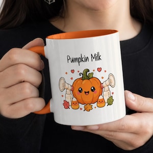 May include: A white ceramic mug with an orange interior and handle, featuring a graphic of a smiling orange pumpkin flanked by two breast pumps and candy corn, with the text Pumpkin Milk printed in black above the illustration.