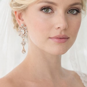 Peut inclure: Boucles d'oreilles de mariée en or rose avec un motif floral et des cristaux pendants. Les boucles d'oreilles sont portées par une personne aux cheveux blonds et un voile. Un bel accessoire pour un mariage ou une occasion spéciale.