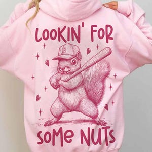 May include: Light pink hoodie with a graphic of a squirrel wearing a baseball cap and holding a bat. The text on the back reads "LOOKIN' FOR SOME NUTS" in a playful font. The design includes hearts and stars.