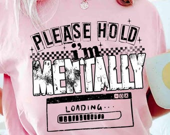 Op de afbeelding: Een lichtroze t-shirt met de tekst "PLEASE HOLD I'M MENTALLY LOADING..." in een zwart-wit ontwerp. Het shirt heeft een ronde hals en korte mouwen. Een witte mok met een gele cirkel wordt in de hand gehouden.
