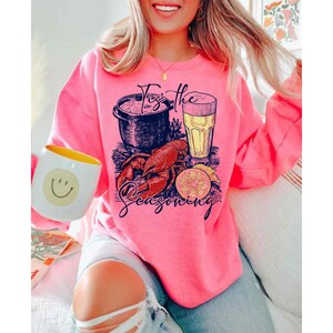 May include: Pink sweatshirt with a graphic of a pot of seafood, a glass of beer, a lobster, and a lemon slice. The text on the sweatshirt says "For the Seasoning".