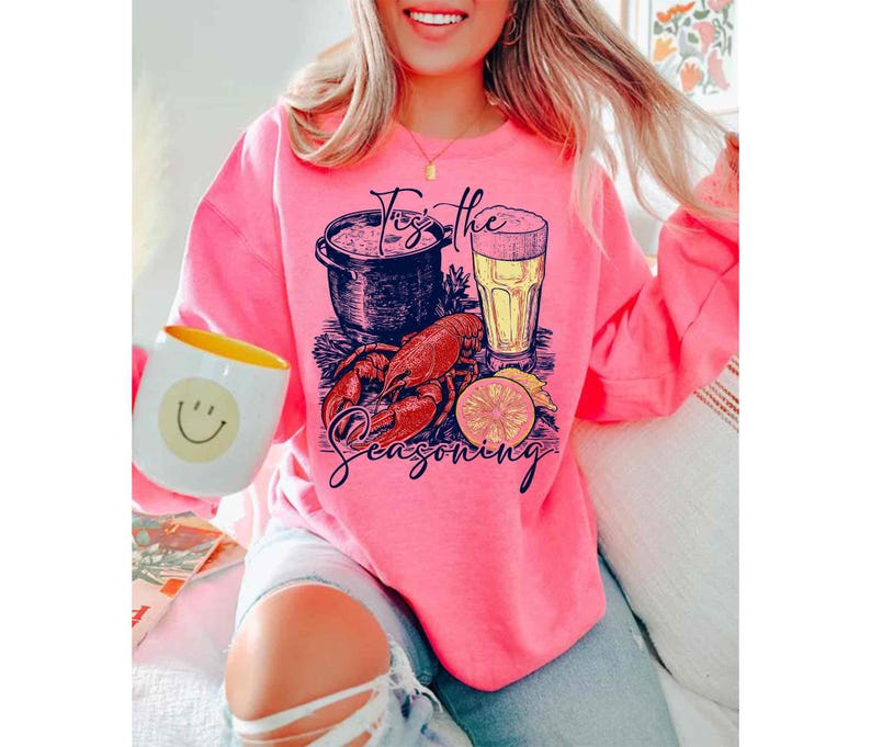 May include: Pink sweatshirt with a graphic of a pot of seafood, a glass of beer, a lobster, and a lemon slice. The text on the sweatshirt says "For the Seasoning".