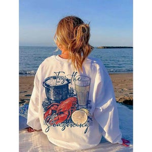 May include: White sweatshirt with a graphic of a pot of food, a glass of beer, a lobster, and a lemon. The text on the sweatshirt says "For the seasoning".