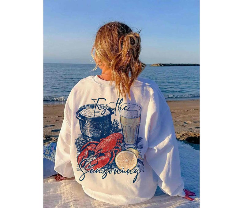 May include: White sweatshirt with a graphic of a pot of food, a glass of beer, a lobster, and a lemon. The text on the sweatshirt says "For the seasoning".