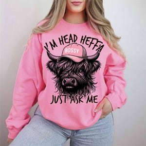 May include: Pink sweatshirt with a black and white illustration of a Highland cow wearing a pink hat that says "Bossy". The text on the sweatshirt reads "I'm Head Heffa Just Ask Me".