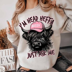 May include: White sweatshirt with a black and white illustration of a Highland cow wearing a pink hat that says "Bossy". The text "I'm Head Heffa Just Ask Me" is printed on the sweatshirt.