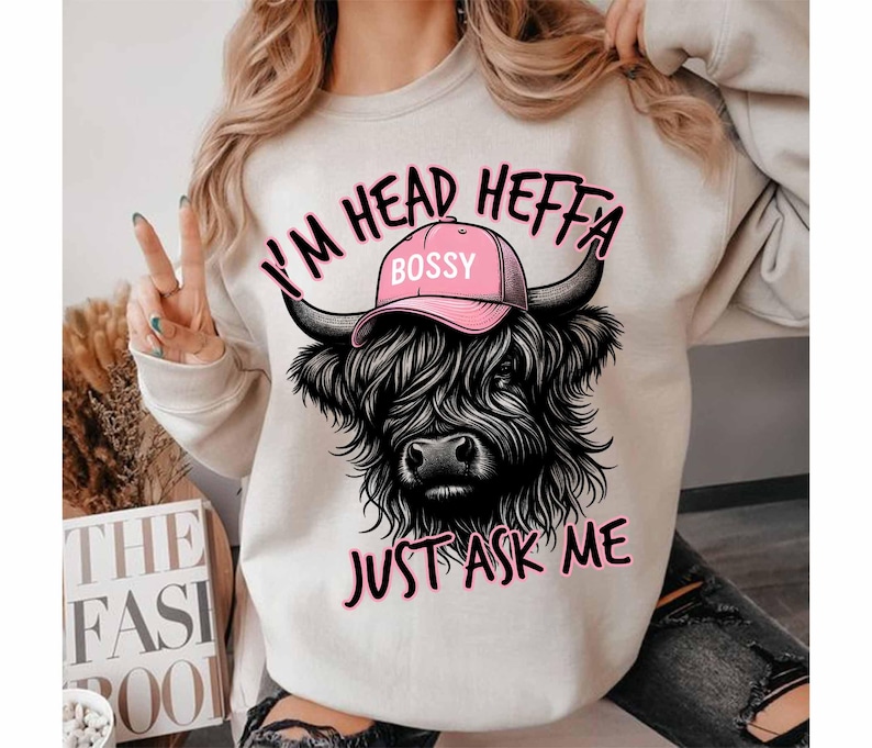 May include: White sweatshirt with a black and white illustration of a Highland cow wearing a pink hat that says "Bossy". The text "I'm Head Heffa Just Ask Me" is printed on the sweatshirt.