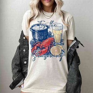 May include: A white t-shirt with a graphic of a pot of seafood, a glass of beer, a lobster, and a lemon slice. The text "Tis the Seasoning" is printed below the graphic.