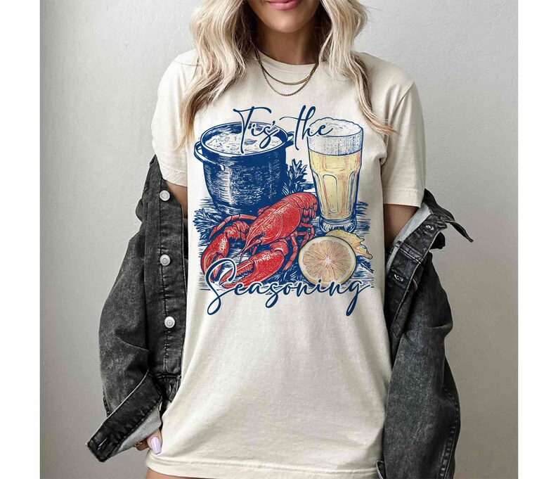 May include: A white t-shirt with a graphic of a pot of seafood, a glass of beer, a lobster, and a lemon slice. The text "Tis the Seasoning" is printed below the graphic.