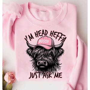 May include: Pink sweatshirt with a black and white illustration of a Highland cow wearing a pink baseball cap that says "Bossy". The text "I'm Head Heffa Just Ask Me" is printed on the sweatshirt.
