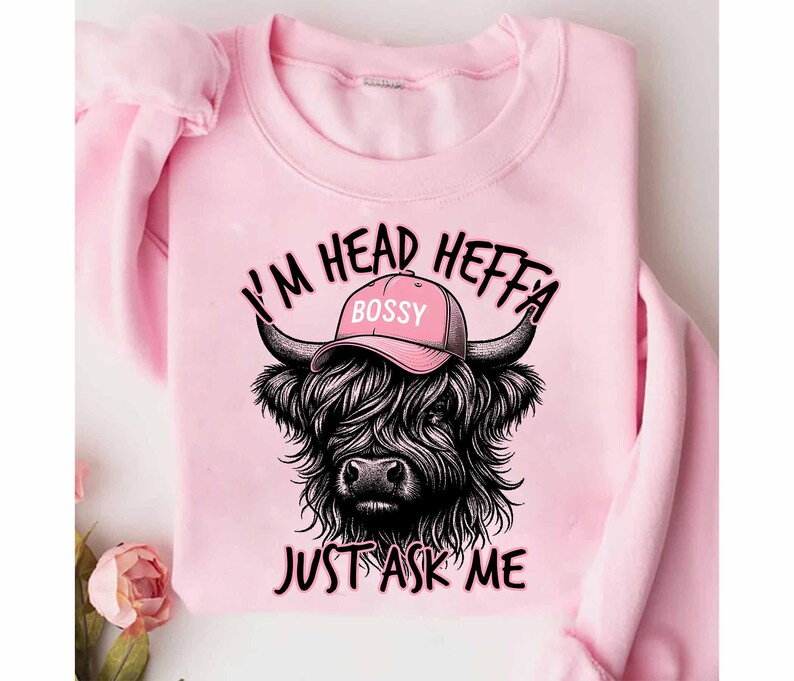 May include: Pink sweatshirt with a black and white illustration of a Highland cow wearing a pink baseball cap that says "Bossy". The text "I'm Head Heffa Just Ask Me" is printed on the sweatshirt.