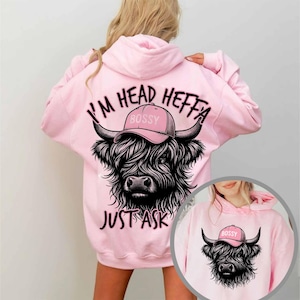 May include: Pink hoodie featuring a black and white Highland cow wearing a pink hat that says "Bossy" and the text "I'm Head Heffa Just Ask".