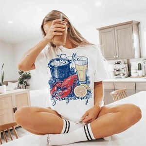 May include: White t-shirt with a graphic of a lobster, a pot of seafood, a glass of beer, and a lemon slice. The text "Toss the Seasoning" is printed on the shirt.