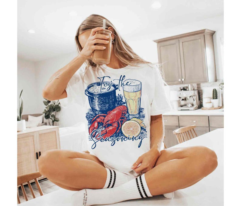 May include: White t-shirt with a graphic of a lobster, a pot of seafood, a glass of beer, and a lemon slice. The text "Toss the Seasoning" is printed on the shirt.