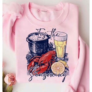 May include: Pink sweatshirt with a graphic design of a pot of seafood, a glass of beer, a lobster, and a lemon. The text "Tis the Seasoning" is printed on the sweatshirt.