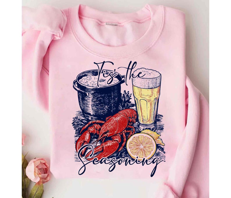 May include: Pink sweatshirt with a graphic design of a pot of seafood, a glass of beer, a lobster, and a lemon. The text "Tis the Seasoning" is printed on the sweatshirt.