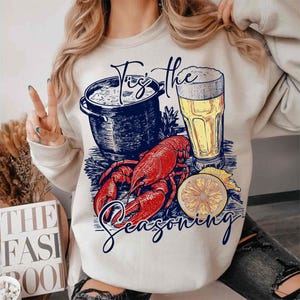 May include: A white sweatshirt with a graphic design of a lobster, a pot of seafood, a glass of beer, and a lemon slice. The text "Tis the Seasoning" is printed on the sweatshirt.