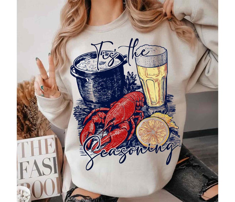May include: A white sweatshirt with a graphic design of a lobster, a pot of seafood, a glass of beer, and a lemon slice. The text "Tis the Seasoning" is printed on the sweatshirt.