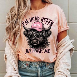 May include: A pink t-shirt with a black and white illustration of a Highland cow wearing a pink hat that says "Bossy". The text on the shirt reads "I'm Head Heffa Just Ask Me".