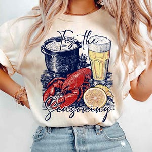 May include: A white t-shirt with a graphic design of a lobster, a pot of seafood, a glass of beer, and a lemon slice. The text "Tis the Seasoning" is printed on the shirt.