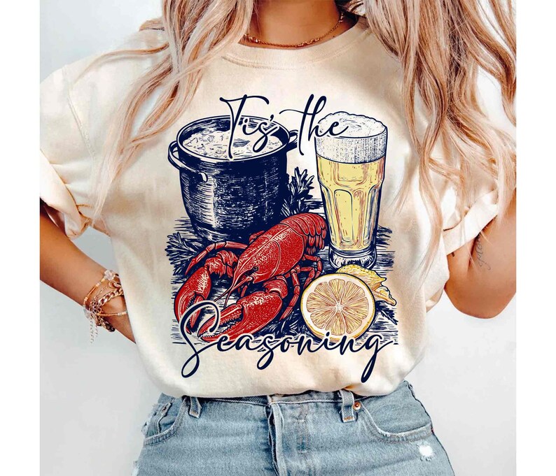 May include: A white t-shirt with a graphic design of a lobster, a pot of seafood, a glass of beer, and a lemon slice. The text "Tis the Seasoning" is printed on the shirt.